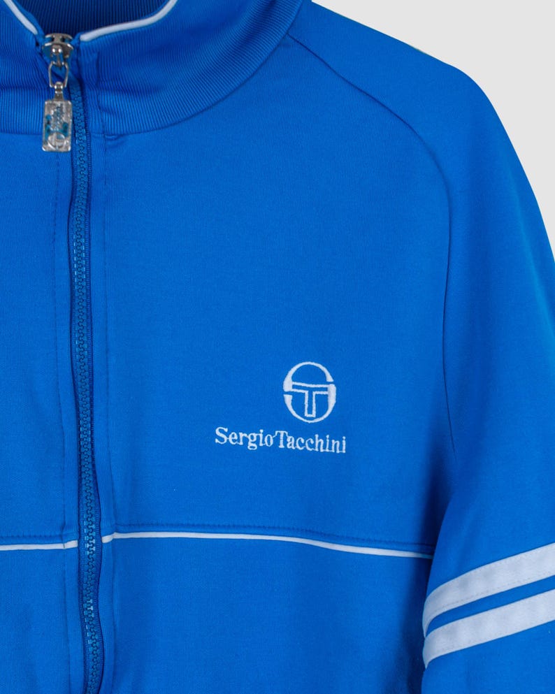 May include: A blue zip-up jacket with white stripes on the sleeves and a white Sergio Tacchini logo on the chest.