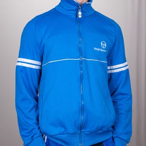 May include: A blue zip-up jacket with white stripes on the sleeves and a white Sergio Tacchini logo on the chest.