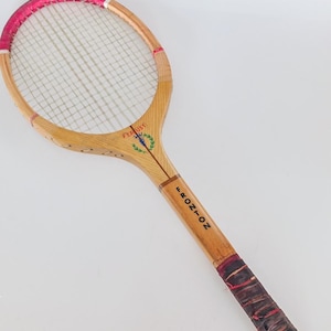 Puede incluir: Raqueta de tenis de madera vintage con marco marrón claro, cuerdas blancas y empuñadura roja y marrón. La raqueta tiene las palabras "FERUST" y "FRONTON" impresas. La parte superior de la raqueta tiene un borde rosa.