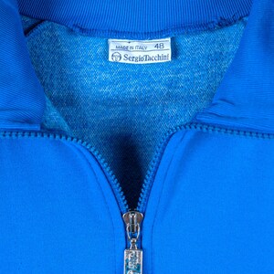 May include: A blue zippered jacket with a silver zipper pull. The label reads "Made in Italy" and "Sergio Tacchini" with the number "48" printed on it.
