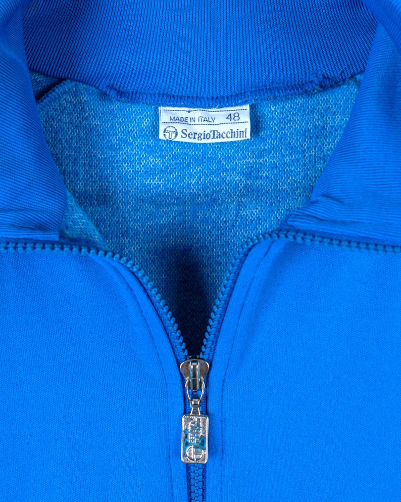 May include: A blue zippered jacket with a silver zipper pull. The label reads "Made in Italy" and "Sergio Tacchini" with the number "48" printed on it.