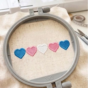 May include: A gray embroidery hoop holds a beige fabric with five embroidered hearts. The hearts are blue, pink, white, pink, and blue. The hoop is labeled "3x3".