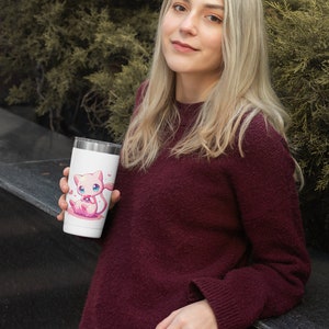 May include: A white stainless steel travel mug with a pink cartoon cat holding a pink ball. The mug has a pink heart design around the cat.
