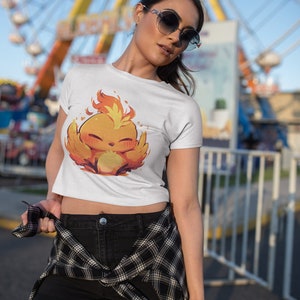 May include: A white crop top with a cartoon image of a yellow and orange bird with flames on its head. The bird is smiling and has its wings spread out. The person wearing the shirt is standing in front of a Ferris wheel.