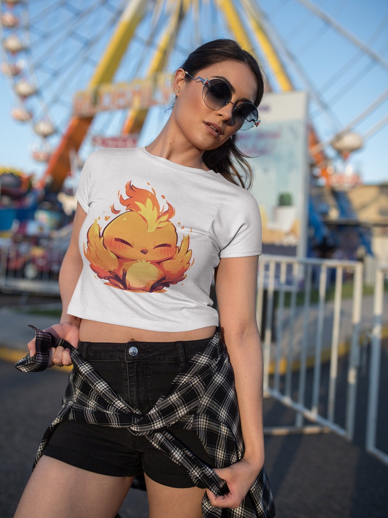 May include: A white crop top with a cartoon image of a yellow and orange bird with flames on its head. The bird is smiling and has its wings spread out. The person wearing the shirt is standing in front of a Ferris wheel.
