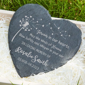 May include: A black heart-shaped slate stone with a dandelion design and the text "Forever in our hearts May the Winds of Heaven Blow Softly and Whisper in your ear IN LOVING MEMORY OF Rosalie Smith 03.1956-06.2023".