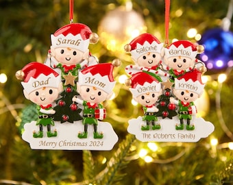 May include: A personalized Christmas ornament featuring a family of elves dressed in green and red, holding a small Christmas tree. The ornament is decorated with the names "Dad", "Sarah", "Mom", "Salome", "Barlow", "Daniel", and "Kristin", along with the text "Merry Christmas 2024" and "The Roberts Family".