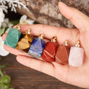Op de afbeelding: Zeven kleine, gefacetteerde edelsteenflesjes met gouden dopjes. De flesjes bevinden zich in een hand, en de edelstenen zijn groen, bruin, blauw, rood, oranje, roze en wit.