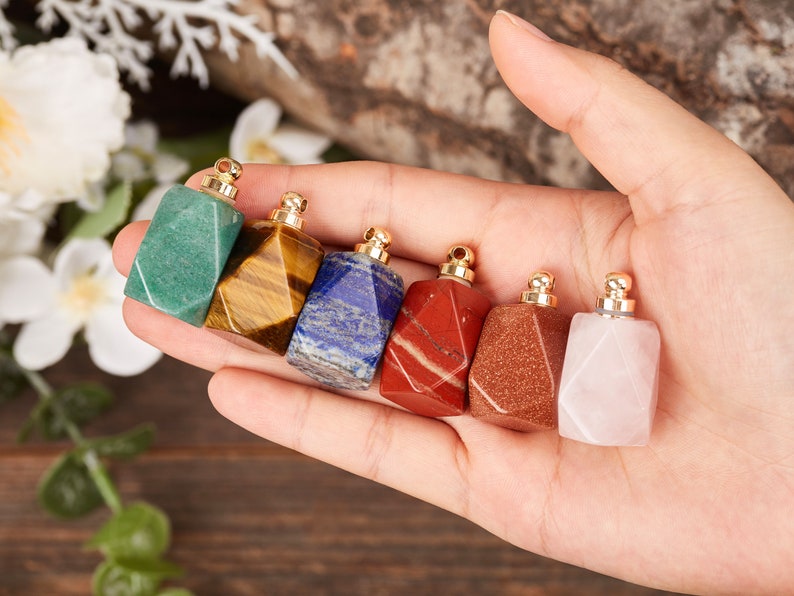 Op de afbeelding: Zeven kleine, gefacetteerde edelsteenflesjes met gouden dopjes. De flesjes bevinden zich in een hand, en de edelstenen zijn groen, bruin, blauw, rood, oranje, roze en wit.