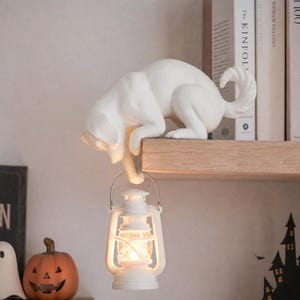 May include: A white cat figurine crouching on the edge of a shelf, holding a small white lantern with a glowing light in its front paw. The cat has a smooth matte finish, a curved tail, and is positioned to hang the lantern below the shelf surface.