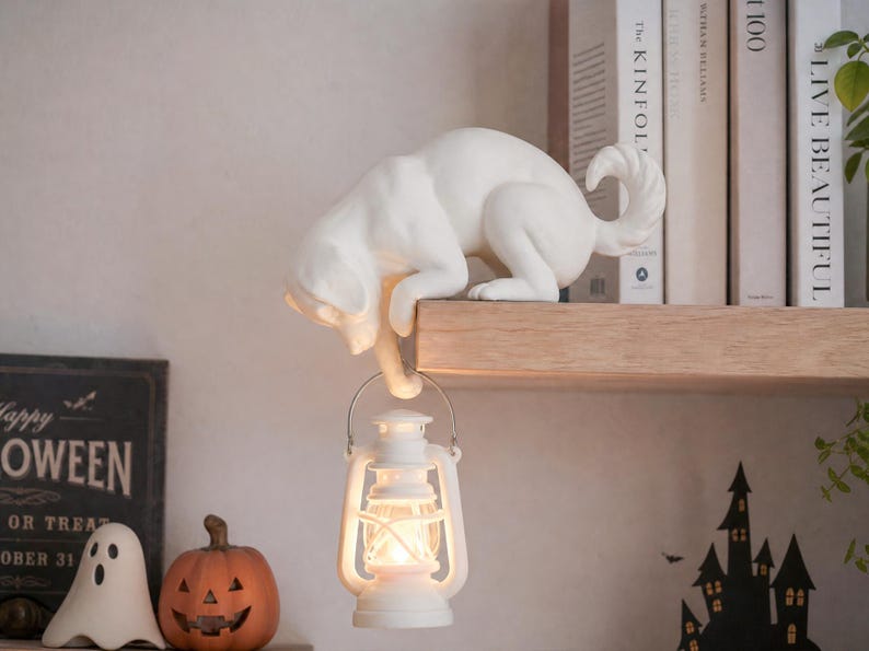 May include: A white cat figurine crouching on the edge of a shelf, holding a small white lantern with a glowing light in its front paw. The cat has a smooth matte finish, a curved tail, and is positioned to hang the lantern below the shelf surface.