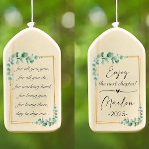 May include: Wooden ornament with a light beige background and a gold border. The ornament has text and a watercolor eucalyptus design. One side reads "for all you give..." and the other side says "Enjoy the next chapter!" with a name and year.