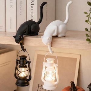 May include: Two cat-shaped figurines, one black and one white, each perched on an edge. Each cat has a long, upright tail and a front paw extending downward to hold a small, battery-operated lantern that emits a warm, glowing light.