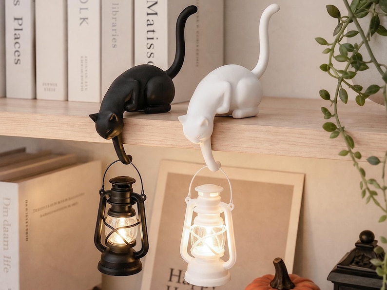 May include: Two cat-shaped figurines, one black and one white, each perched on an edge. Each cat has a long, upright tail and a front paw extending downward to hold a small, battery-operated lantern that emits a warm, glowing light.