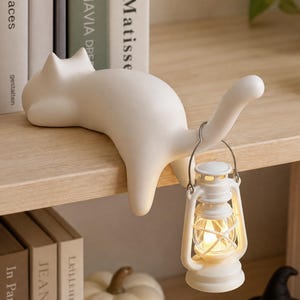 May include: A matte white cat figurine lying on a shelf edge with its tail curved upward. A small, white, battery-operated lantern with a clear glass globe and a metal handle hangs from the cat's tail, emitting a warm, soft, glowing light.