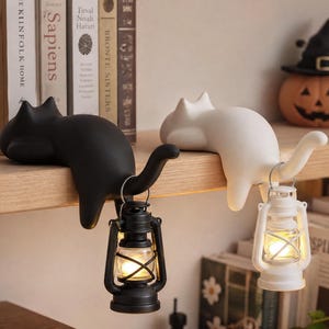 May include: Two cat-shaped shelf sitters, one matte black and one matte white, each featuring a curved tail that acts as a hook. A small black lantern hangs from the black cat's tail, and a small white lantern hangs from the white cat's tail.