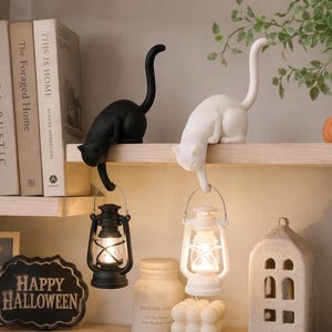May include: Two cat-shaped figurines, one black and one white, are perched on a shelf. Each cat has a long, upright tail and holds a small, battery-operated lantern suspended from its front paw. The lanterns emit a warm, glowing light.