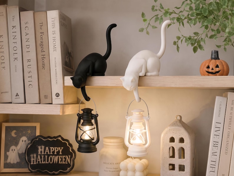May include: Two cat-shaped figurines, one black and one white, are perched on a shelf. Each cat has a long, upright tail and holds a small, battery-operated lantern suspended from its front paw. The lanterns emit a warm, glowing light.