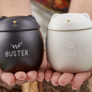 Puede incluir: Dos urnas con forma de gato, una negra y otra blanca, se sostienen en manos ahuecadas. La urna negra tiene el nombre "BUSTER" y un diseño de ramita, mientras que la urna blanca tiene "MOLLY" y una huella de pata con alas. Ambas tienen detalles dorados.
