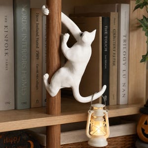 May include: A white cat figurine climbing a vertical wooden pole. The cat has a curved tail that acts as a hook, from which a small, battery-operated lantern with a warm, glowing light is suspended. The figurine is positioned in front of books.