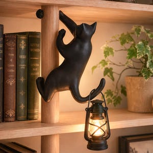 May include: A matte black cat figurine climbing a vertical wooden pole, with its tail curved to hold a small, lit black lantern. The cat is positioned on a shelf next to several books, a small pumpkin, a potted plant, and a skull decoration.