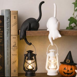 May include: Two cat-shaped figurines, one black and one white, are perched on a shelf. Each cat holds a small, matching colored lantern by its handle. The lanterns emit a warm, glowing light, and the cats have their tails curved upward.