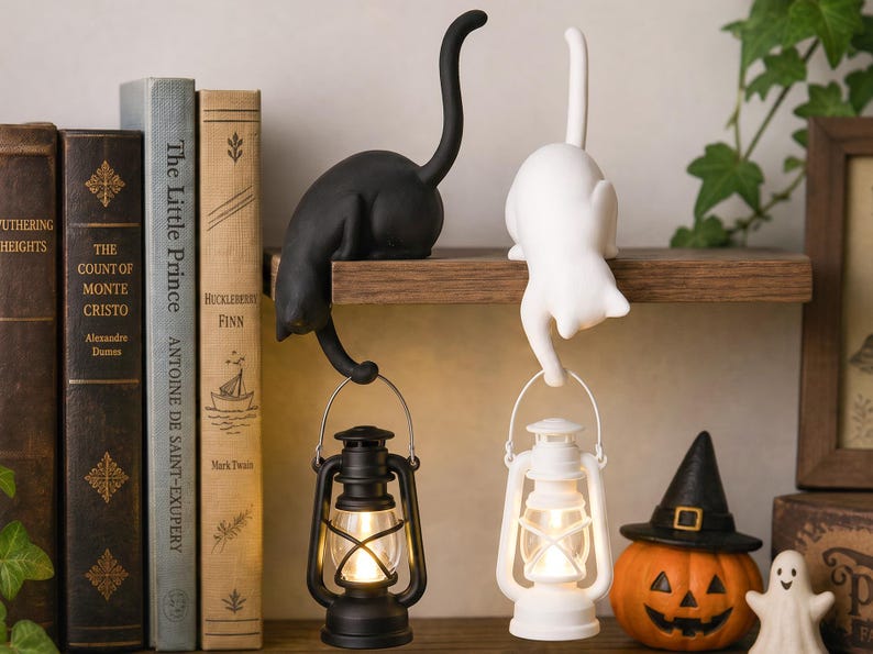 May include: Two cat-shaped figurines, one black and one white, are perched on a shelf. Each cat holds a small, matching colored lantern by its handle. The lanterns emit a warm, glowing light, and the cats have their tails curved upward.
