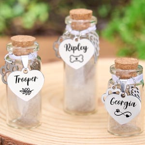 May include: Three small glass bottles with cork stoppers, each with a heart-shaped tag. The tags are engraved with names and pet-themed designs. The bottles are filled with a light-colored material and are displayed on a wooden surface.