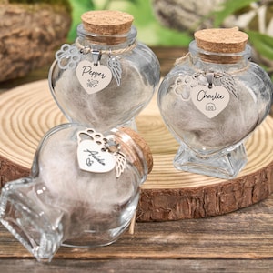 Może przedstawiać: Trzy przezroczyste, szklane słoiczki w kształcie serca z korkowymi zatyczkami, każdy ze srebrnym urokiem i etykietą z imieniem. Słoiki stoją na drewnianym plastrze, wypełnione miękkim, jasnym materiałem, prawdopodobnie do upamiętniania zwierząt.