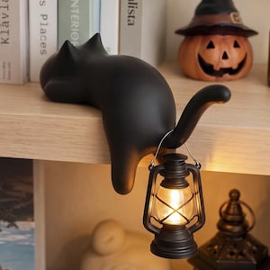 May include: A matte black cat figurine lying on a shelf edge with its tail curved upward. A small, battery-operated black lantern with a warm glowing light hangs from the cat's tail. The cat has pointed ears and a smooth, solid-colored surface.