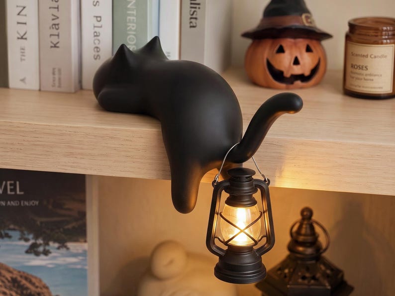 May include: A matte black cat figurine lying on a shelf edge with its tail curved upward. A small, battery-operated black lantern with a warm glowing light hangs from the cat's tail. The cat has pointed ears and a smooth, solid-colored surface.