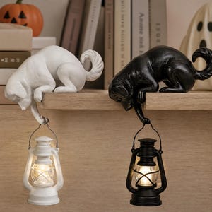 May include: Two dog figurines, one white and one black, are perched on a ledge. Each dog holds a small, lit lantern suspended from its front paw. The lanterns feature a classic design with a wire handle and a glowing light source inside the glass.