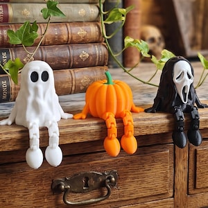 Peut inclure: Trois figurines articulées assises sur une étagère, représentant un fantôme blanc, une citrouille orange et un personnage masqué noir. Chaque figurine possède des membres flexibles et est conçue pour s'asseoir sur le bord d'une surface.