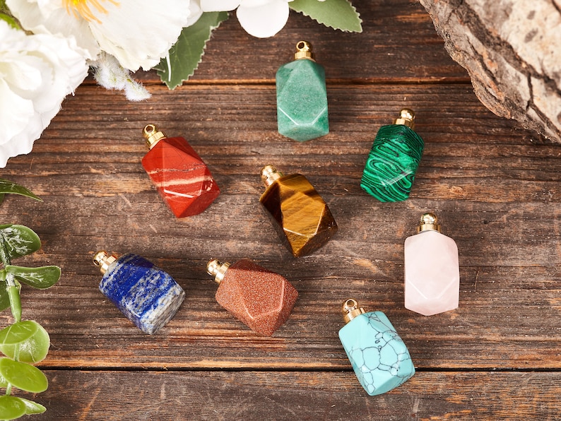 Op de afbeelding: Acht kleine edelsteenflesjes met gouden dopjes. De flesjes zijn gemaakt van verschillende stenen, waaronder rood, groen, bruin, blauw, roze en turquoise.