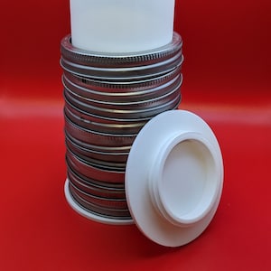 May include: A stack of white plastic jar lids with a white plastic cylinder on top. The lids are stacked on a red surface.
