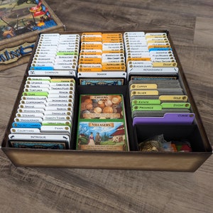 May include: A board game organizer with labeled card dividers. The cards are arranged in a brown tray, with various categories like "Copper," "Silver," and "Province." The game box is visible in the background.