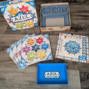 May include: The board game "Azul: Summer Pavilion" is displayed, featuring a box, game boards, and tiles. The box has a colorful design with the game's title. The game boards and tiles have geometric patterns in various colors, including blue, red, and yellow. A blue tray is also visible.