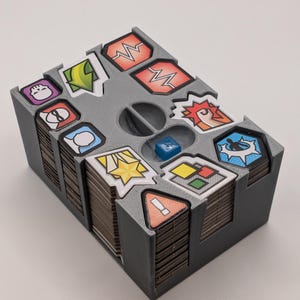 May include: A gray plastic organizer with compartments for storing small game pieces. The organizer has a variety of colorful stickers with different symbols on them, including a fist, a lightning bolt, a star, and an exclamation point.