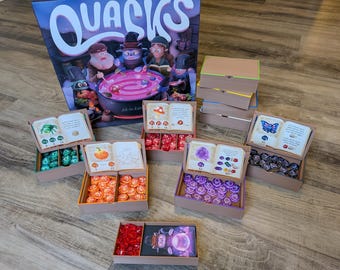 May include: A board game titled "Quacks" with various components. The game includes a box, open boxes with colorful game pieces, and cards. The game pieces are in shades of green, orange, red, purple, and black.