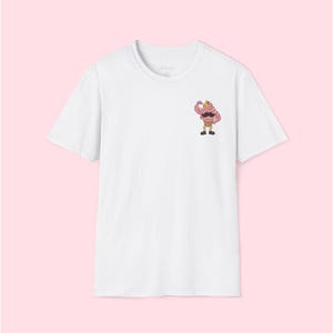 May include: White t-shirt featuring a cartoon ice cream cone character with a mustache and flexing arm. The ice cream cone is pink and brown, with black shoes and a yellow background. The shirt has a crew neck.
