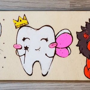 May include: A wooden plaque featuring cartoon illustrations of a mouse with a pink bow, a mouse with a tooth, a tooth with a crown and fairy wings, a red lion, and a mouse with a blue bow and a gold coin. The illustrations are in a cartoon style.