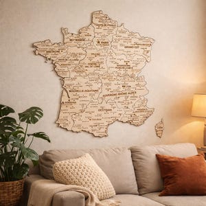 Może przedstawiać: Drewniana mapa Francji, wycinana laserowo z szczegółowymi granicami regionalnymi i nazwami miast. Mapa jest zamontowana na ścianie nad sofą z poduszkami i rośliną. Mapa zawiera tekst: BRETAGNE, PARIS i BORDEAUX. Wymiary są w centymetrach.