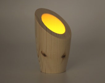 May include: A wooden candle holder with a warm yellow glow emanating from the top. The candle holder is made of light-colored wood and has a cylindrical shape with a slanted top. The wood grain is visible, adding to its natural aesthetic.