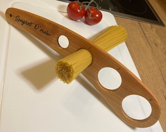 May include: Wooden spaghetti measuring tool with the text "Spaghetti O'meter". The tool has four holes for measuring pasta portions, with spaghetti in the second hole. Two red tomatoes sit nearby.