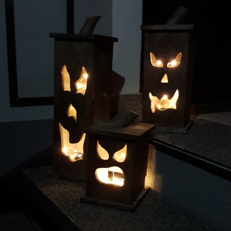 May include: Three wooden Halloween lanterns with carved faces and glowing lights. The lanterns are rectangular with a small chimney-like top. The faces have cut-out eyes, noses, and mouths, creating a spooky effect. The lanterns are different sizes.