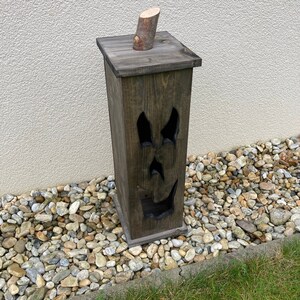 May include: A dark stained wooden Halloween decoration shaped like a tall, rectangular box. The box has a carved face with eye and mouth cutouts. A small wooden log piece is attached to the top. The decoration is placed on a bed of small stones.