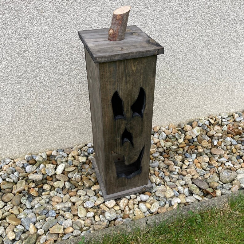 May include: A dark stained wooden Halloween decoration shaped like a tall, rectangular box. The box has a carved face with eye and mouth cutouts. A small wooden log piece is attached to the top. The decoration is placed on a bed of small stones.