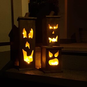 May include: Three wooden Halloween lanterns with carved jack-o'-lantern faces. Each lantern has a rectangular shape and is lit from within by small, warm-toned lights. The lanterns are arranged at different heights, creating a spooky display.