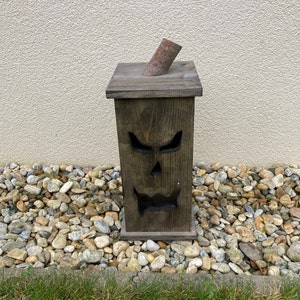 May include: A dark brown wooden Halloween decoration shaped like a jack-o'-lantern. The box has a carved face with triangular eyes, a nose, and a jagged mouth. A cylindrical piece of wood is angled out of the top. The decoration is placed on a bed of rocks.
