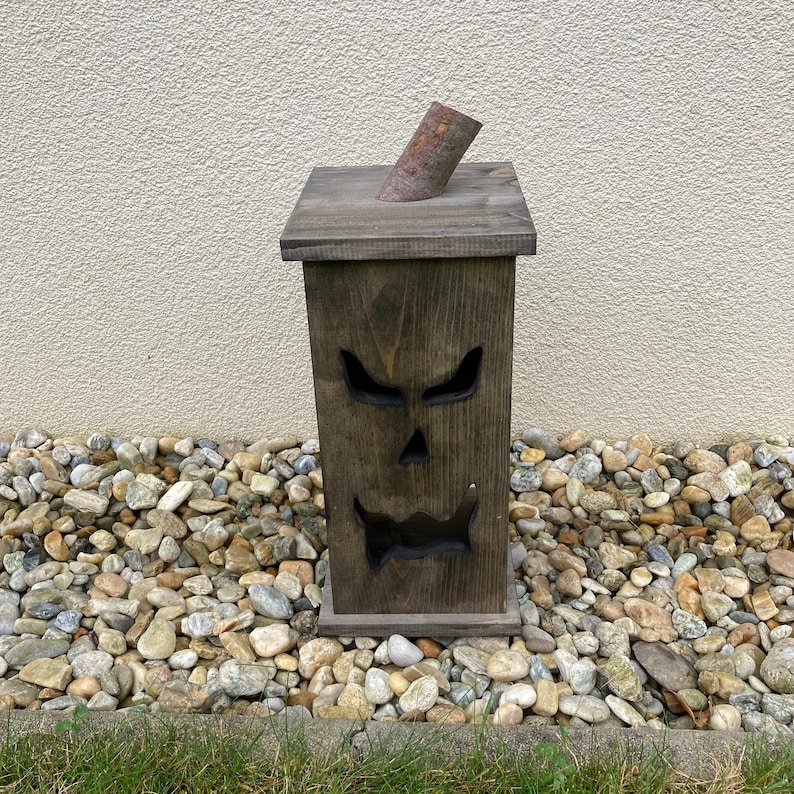 May include: A dark brown wooden Halloween decoration shaped like a jack-o'-lantern. The box has a carved face with triangular eyes, a nose, and a jagged mouth. A cylindrical piece of wood is angled out of the top. The decoration is placed on a bed of rocks.
